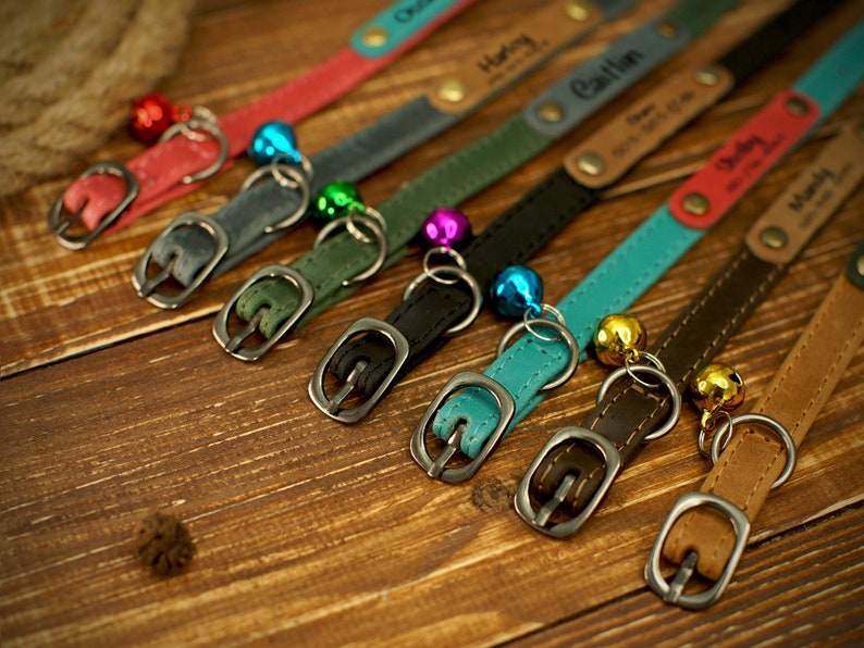 May include: A collection of personalized pet collars in various colors, including red, gray, green, black, teal, brown, and tan. Each collar features a metal buckle, a small bell, and a name tag. The collars are arranged on a wooden surface.