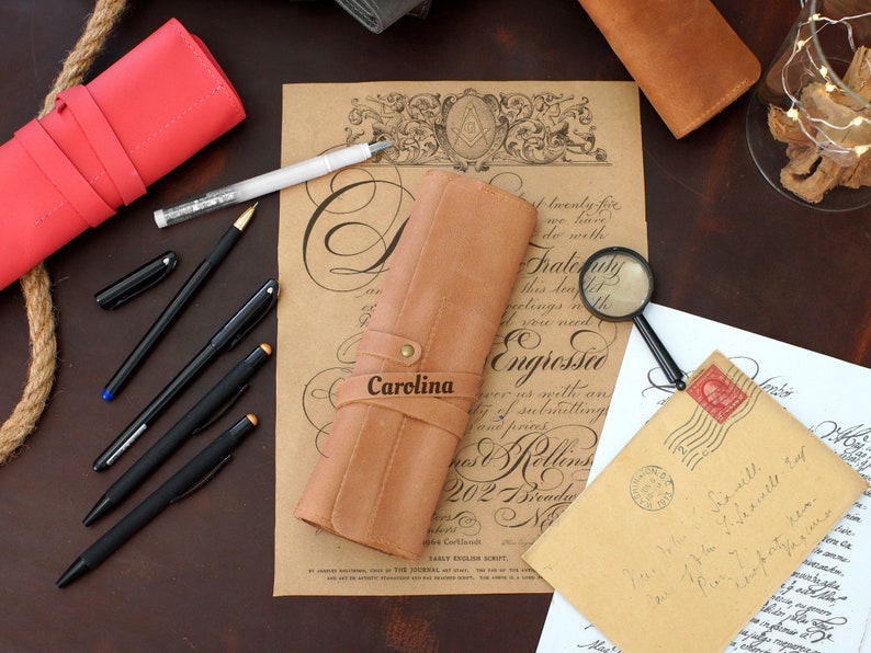 May include: A brown leather pen roll with the name "Carolina" embossed on the front. The roll is open and has several pens inside. The roll is sitting on a table with other writing supplies, including a magnifying glass, a letter, and a red leather roll.