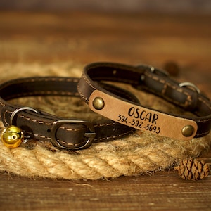 Puede incluir: Dos collares de perro de cuero marr&oacute;n con una campana en uno. El collar m&aacute;s grande tiene el nombre "OSCAR" y el n&uacute;mero de tel&eacute;fono "394-392-8693" grabado en &eacute;l.