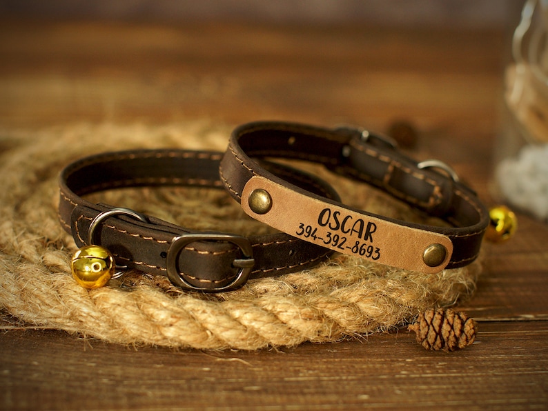 Puede incluir: Dos collares de perro de cuero marr&oacute;n con una campana en uno. El collar m&aacute;s grande tiene el nombre "OSCAR" y el n&uacute;mero de tel&eacute;fono "394-392-8693" grabado en &eacute;l.