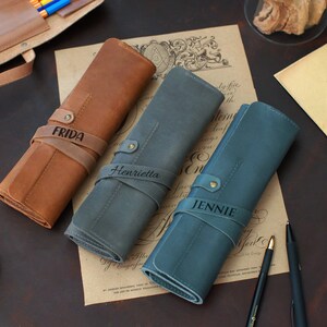 May include: Three personalized leather pencil cases in brown, gray, and blue. The cases are rolled up and have leather straps with the names "Frida", "Henrietta", and "Jennie" engraved on them.