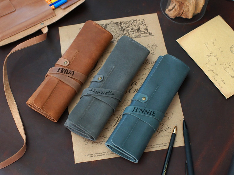 May include: Three personalized leather pencil cases in brown, gray, and blue. The cases are rolled up and have leather straps with the names "Frida", "Henrietta", and "Jennie" engraved on them.