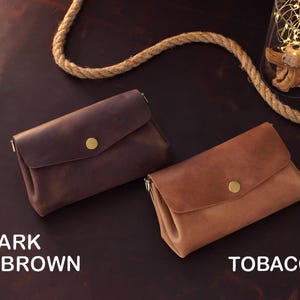 Op de afbeelding: Twee leren clutch portemonnees in donkerbruine en tabakskleuren, elk met een klepsluiting en een goudkleurige knoop. De teksten "DARK BROWN" en "TOBACCO" staan onder de portemonnees. De portemonnees liggen op een donkerbruin oppervlak.