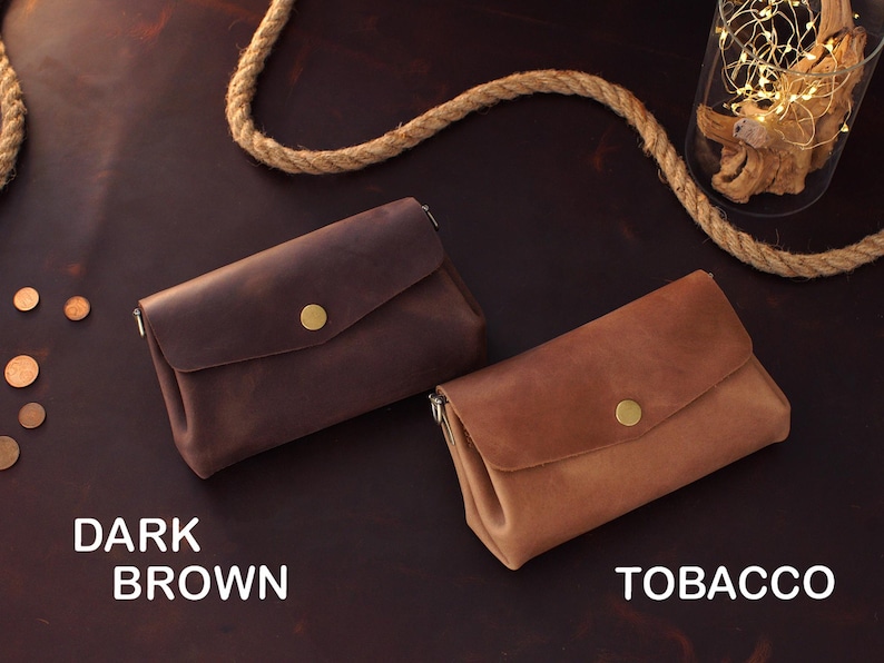 Op de afbeelding: Twee leren clutch portemonnees in donkerbruine en tabakskleuren, elk met een klepsluiting en een goudkleurige knoop. De teksten "DARK BROWN" en "TOBACCO" staan onder de portemonnees. De portemonnees liggen op een donkerbruin oppervlak.