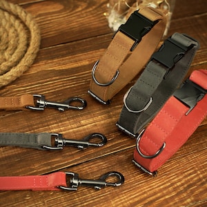May include: A selection of dog collars and leashes in various colors, including tan, gray, and red. The collars feature black buckles and metal D-rings. The leashes have matching colors and metal clasps. The items are arranged on a wooden surface.