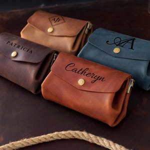 May include: An assortment of personalized leather pouches in various colors: brown, tan, and blue. Each pouch features a flap closure with a gold-colored button and a zipper. The pouches are embossed with names or initials.