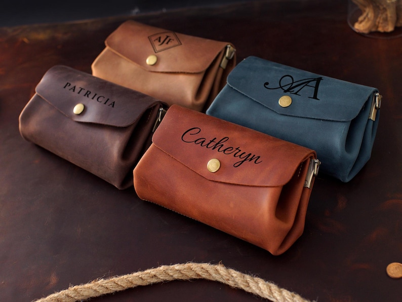 May include: An assortment of personalized leather pouches in various colors: brown, tan, and blue. Each pouch features a flap closure with a gold-colored button and a zipper. The pouches are embossed with names or initials.
