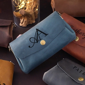 Op de afbeelding: Een verzameling gepersonaliseerde leren portemonnees in verschillende kleuren, waaronder blauw, bruin en grijs. Elke portemonnee heeft een klepsluiting met een goudkleurige knoop en een ritssluiting. Sommige portemonnees zijn gegraveerd met initialen of namen.