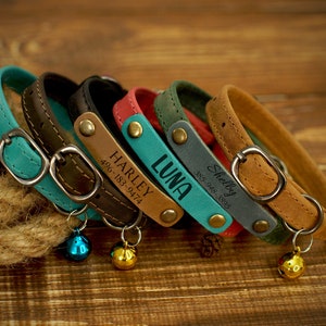 May include: A collection of personalized leather cat collars in various colors, including teal, brown, black, red, green, and tan. Each collar features a metal buckle, a name tag with a name and phone number, and a small bell.