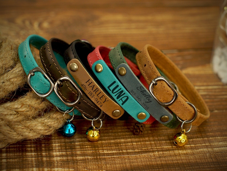 May include: A collection of personalized leather cat collars in various colors, including teal, brown, black, red, green, and tan. Each collar features a metal buckle, a name tag with a name and phone number, and a small bell.