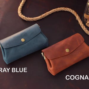 Op de afbeelding: Twee leren clutch portemonnees, &eacute;&eacute;n grijsblauw en &eacute;&eacute;n cognac, worden getoond op een donkerbruin oppervlak. Elke portemonnee heeft een klepsluiting met een goudkleurige knoop. De teksten "GRAY BLUE" en "COGNAC" zijn zichtbaar.