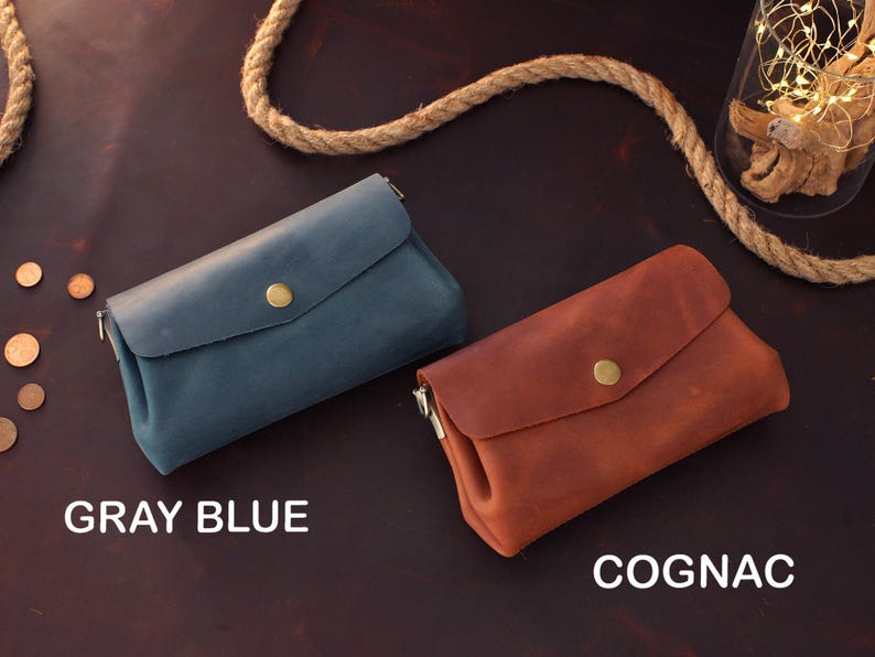 Op de afbeelding: Twee leren clutch portemonnees, &eacute;&eacute;n grijsblauw en &eacute;&eacute;n cognac, worden getoond op een donkerbruin oppervlak. Elke portemonnee heeft een klepsluiting met een goudkleurige knoop. De teksten "GRAY BLUE" en "COGNAC" zijn zichtbaar.