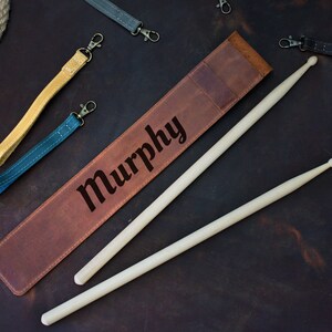 Op de afbeelding: Een bruine leren drumstickhouder met de naam "Murphy" erop gegraveerd. Twee houten drumsticks liggen op de houder.