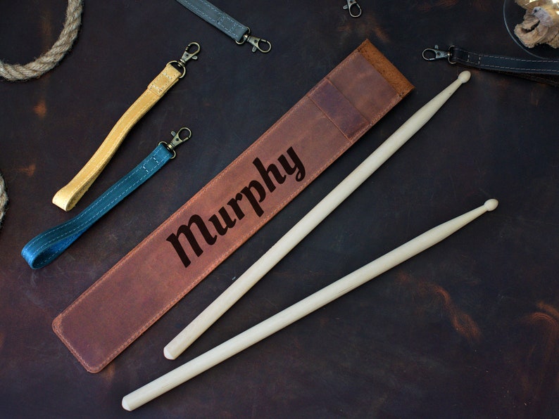 Op de afbeelding: Een bruine leren drumstickhouder met de naam "Murphy" erop gegraveerd. Twee houten drumsticks liggen op de houder.
