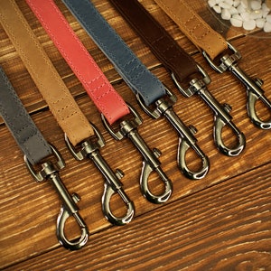 May include: A collection of six leather dog leashes in various colors, including gray, tan, coral, blue, brown, and light brown. Each leash features a silver-colored metal clasp and is displayed on a wooden surface.