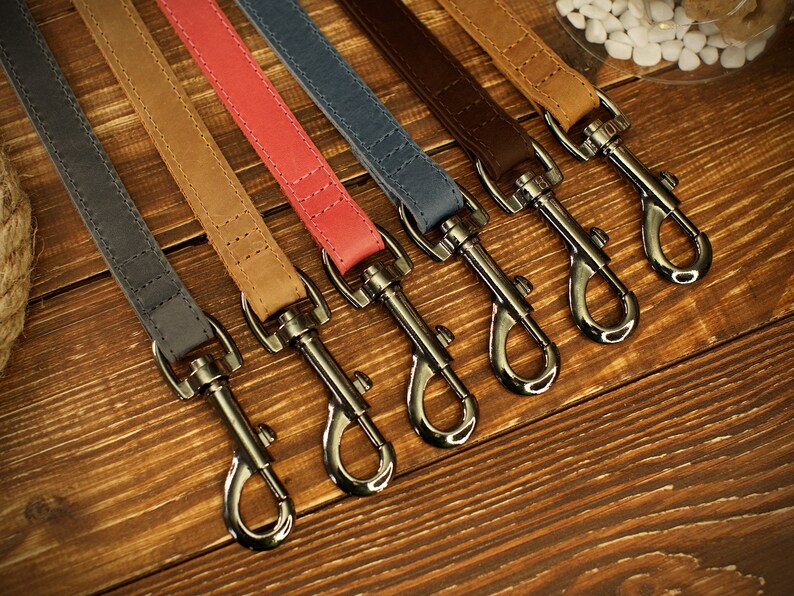 May include: A collection of six leather dog leashes in various colors, including gray, tan, coral, blue, brown, and light brown. Each leash features a silver-colored metal clasp and is displayed on a wooden surface.