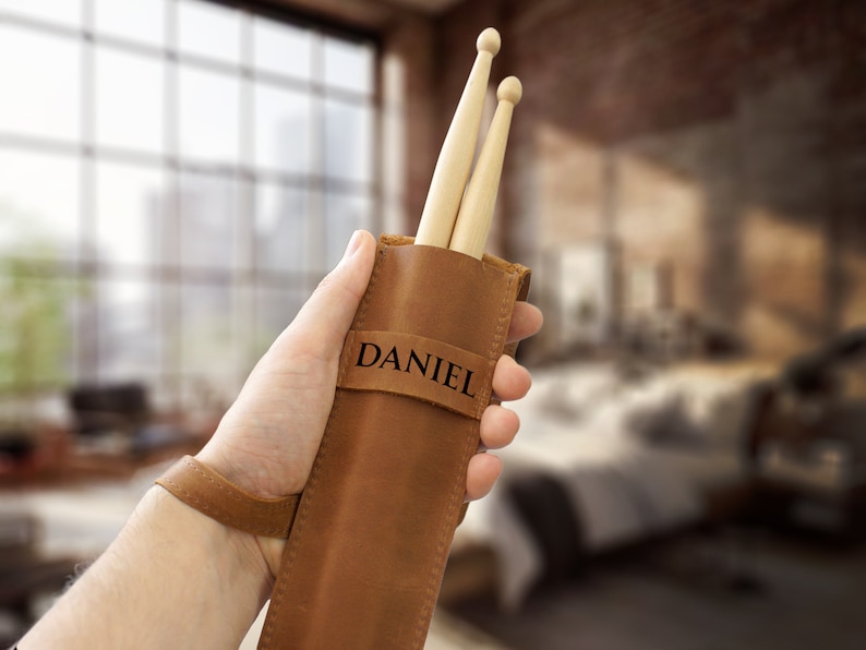 Op de afbeelding: Een bruine leren drumstickhouder met een band en de naam "DANIEL" gegraveerd op de voorkant. Twee houten drumsticks zitten in de houder.
