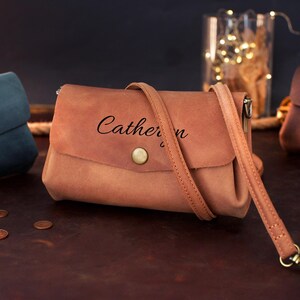 May include: A light brown leather crossbody bag with a flap closure and a long strap. The bag has the name "Catherine" printed on the front. Other bags in blue and brown are in the background.