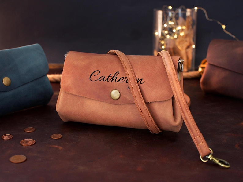 May include: A light brown leather crossbody bag with a flap closure and a long strap. The bag has the name "Catherine" printed on the front. Other bags in blue and brown are in the background.