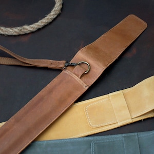 Op de afbeelding: Twee leren pennenkokers, een bruin en een beige, met een bruine leren band en een metalen ring. De kokers liggen op een bruin oppervlak.