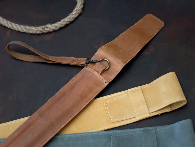 Op de afbeelding: Twee leren pennenkokers, een bruin en een beige, met een bruine leren band en een metalen ring. De kokers liggen op een bruin oppervlak.