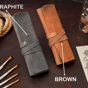 May include: Two leather pencil cases, one in brown and one in graphite gray. Both cases have a strap closure and are rolled up. The cases are on a wooden surface with paint brushes.