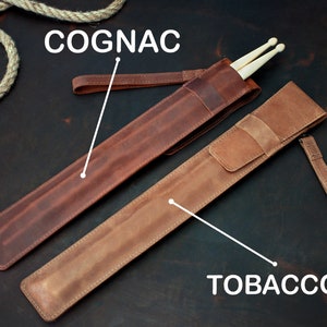 Op de afbeelding: Twee leren drumstick-hoezen, een in cognac en een in tabak. Beide hoezen hebben een band.