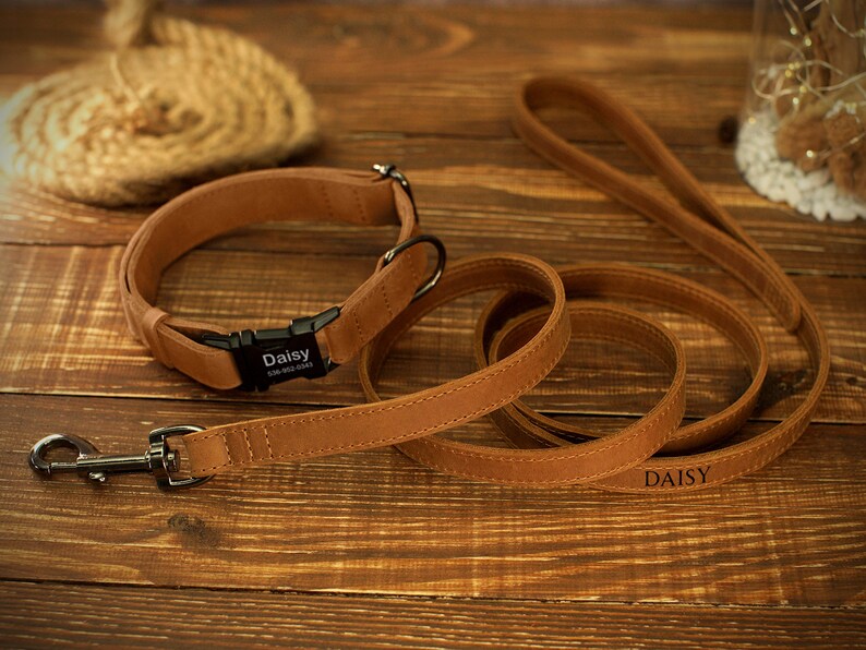 May include: A tan leather dog collar and matching leash set. The collar has a black buckle with the name "Daisy" and a phone number. The leash also has the name "Daisy" engraved on it. The set is on a wooden surface.