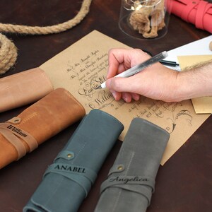 May include: Four personalized leather pen rolls in brown, tan, blue, and gray. The rolls are rolled up and have a strap with a name engraved on each. The names are Bessie, William, Anabel, and Angelica.