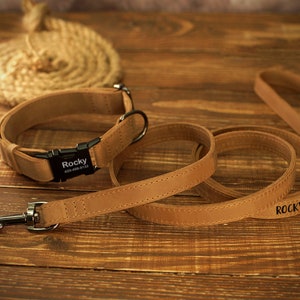 May include: A tan leather dog collar and matching leash set. The collar has a black buckle with the name "Rocky" and a phone number. The leash also has the name "Rocky" printed on it. The set is on a wooden surface.