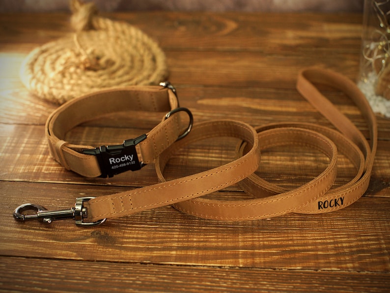 May include: A tan leather dog collar and matching leash set. The collar has a black buckle with the name "Rocky" and a phone number. The leash also has the name "Rocky" printed on it. The set is on a wooden surface.