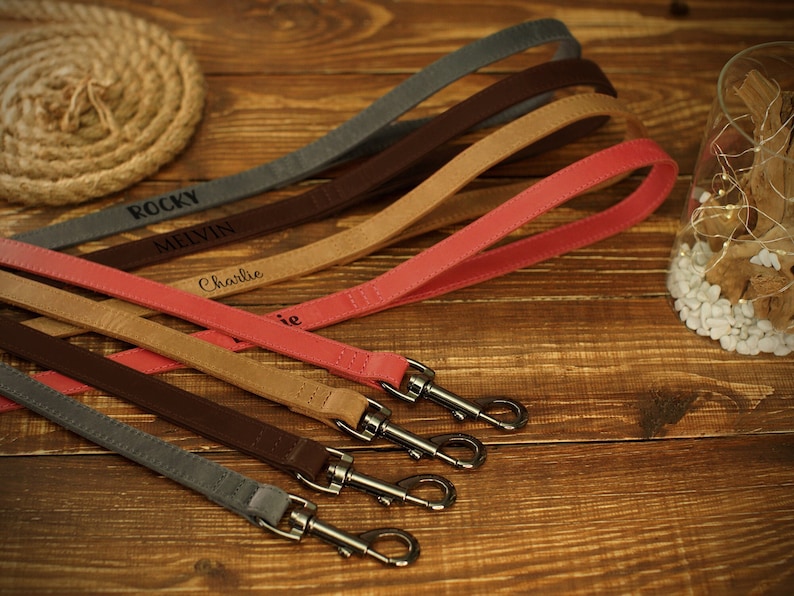 May include: A collection of personalized dog leashes in various colors, including gray, brown, tan, and pink. Each leash features a silver-colored clasp and is engraved with a dog's name. The leashes are arranged on a wooden surface.