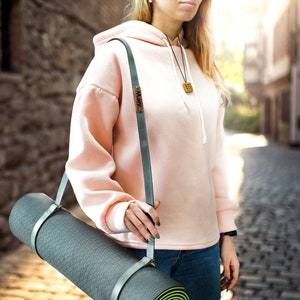 May include: A person carrying a gray yoga mat with a light gray strap. The strap has a small metal tag with the word "Mary" on it. The person is wearing a pink hoodie and blue jeans.