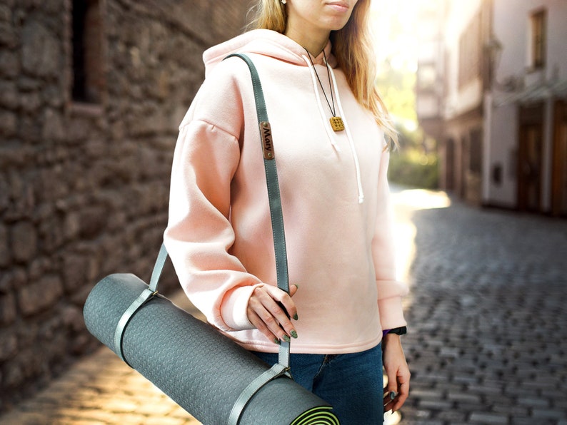 May include: A person carrying a gray yoga mat with a light gray strap. The strap has a small metal tag with the word "Mary" on it. The person is wearing a pink hoodie and blue jeans.