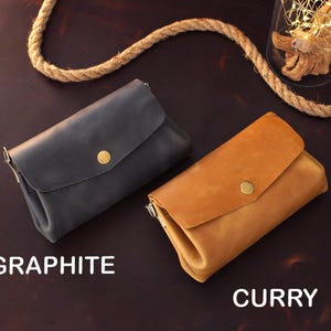 Op de afbeelding: Twee leren portemonnees, &eacute;&eacute;n grafiet en &eacute;&eacute;n curry, met een klepsluiting en een goudkleurige knoop. De portemonnees worden gepresenteerd op een donkerbruin oppervlak. Het woord "GRAPHITE" staat onder de grafietkleurige portemonnee en "CURRY" onder de currykleurige portemonnee.