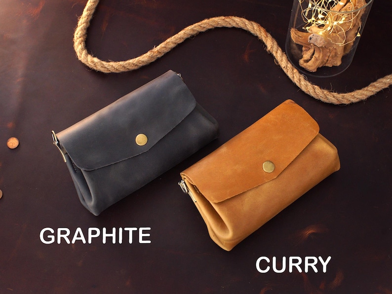 Op de afbeelding: Twee leren portemonnees, &eacute;&eacute;n grafiet en &eacute;&eacute;n curry, met een klepsluiting en een goudkleurige knoop. De portemonnees worden gepresenteerd op een donkerbruin oppervlak. Het woord "GRAPHITE" staat onder de grafietkleurige portemonnee en "CURRY" onder de currykleurige portemonnee.