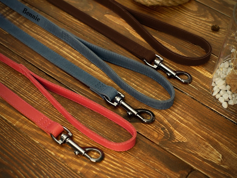 May include: A selection of dog leashes in various colors, including red, blue, and brown. Each leash features a metal clasp and a loop handle. One leash is personalized with the name "Bonnie". The leashes are arranged on a wooden surface.