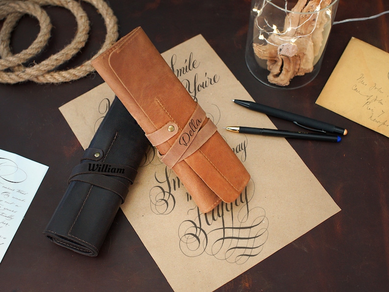 May include: Two personalized leather pen rolls, one brown and one black, with the names "Della" and "William" embossed on them. The rolls are lying on a piece of paper with calligraphy writing.