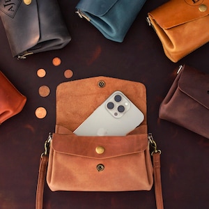 May include: A collection of leather phone crossbody bags in various colors, including brown, tan, and dark gray. One bag is open, revealing a smartphone. Each bag has a snap closure and a gold-colored button.
