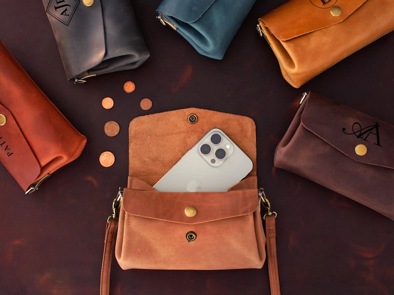 May include: A collection of leather phone crossbody bags in various colors, including brown, tan, and dark gray. One bag is open, revealing a smartphone. Each bag has a snap closure and a gold-colored button.