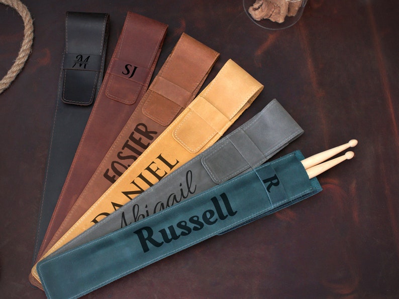Op de afbeelding: Zes gepersonaliseerde leren drumstick-houders in verschillende kleuren, waaronder bruin, beige, zwart en grijs. De houders liggen op een bruin oppervlak en hebben verschillende namen, waaronder "M", "SJ", "Foster", "Daniel", "Abigail" en "Russell".