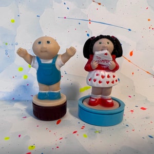 Peut inclure: Deux figurines Cabbage Patch Kids, l'une portant une salopette bleue et l'autre une robe rouge à cœurs blancs. Les figurines sont sur un fond blanc avec des éclaboussures de peinture colorées.