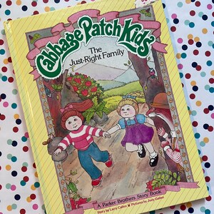 May include: A yellow book with a pink and green Cabbage Patch Kids logo. The title is "The Just-Right Family". The book is a Parker Brothers Story Book with a story by Larry Callen and pictures by Judy Gallen.