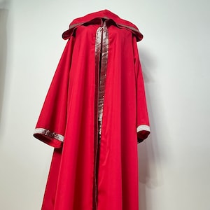 May include: A floor-length red hooded cloak featuring a wide hood and long sleeves. The garment is accented with a decorative metallic gold trim along the front opening, the edges of the hood, and the cuffs, displaying a repeating geometric pattern.