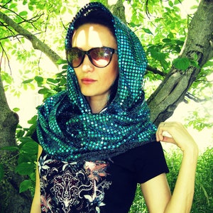 May include: A woman wearing a teal sequined scarf with a black shirt. The scarf is wrapped around her head and neck. She is wearing sunglasses and has a neutral expression.