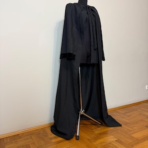 May include: A long, black cloak with a high collar and velvet trim on the sleeves is displayed on a mannequin. The cloak is floor-length and has a split front, revealing the garment underneath. The background is a neutral wall and wooden floor.