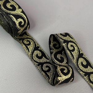 May include: A roll of black ribbon with a gold, ornate, swirling pattern. The ribbon is partially unrolled, revealing the detailed design. The ribbon is likely used for decorative purposes, such as embellishing clothing or crafts.