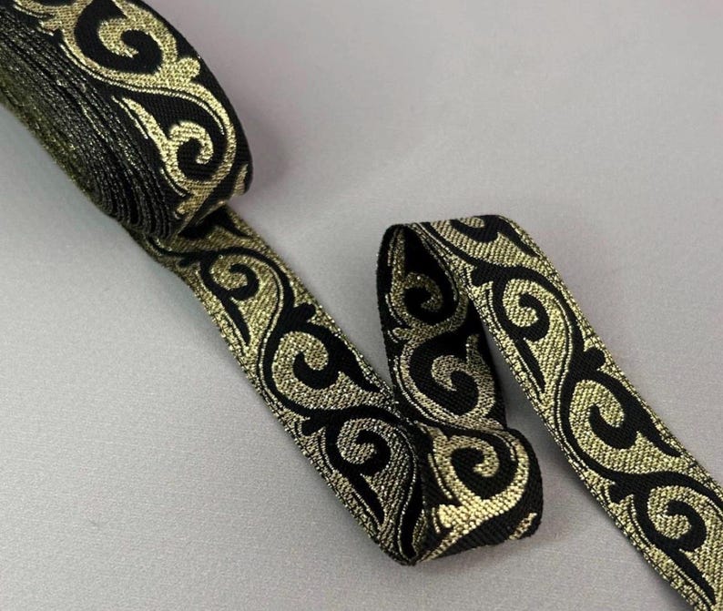 May include: A roll of black ribbon with a gold, ornate, swirling pattern. The ribbon is partially unrolled, revealing the detailed design. The ribbon is likely used for decorative purposes, such as embellishing clothing or crafts.