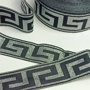 May include: Black and silver decorative ribbon with a Greek key pattern. The ribbon is rolled and unrolled, showcasing the repeating geometric design. The ribbon's metallic sheen adds a touch of elegance to the overall appearance.