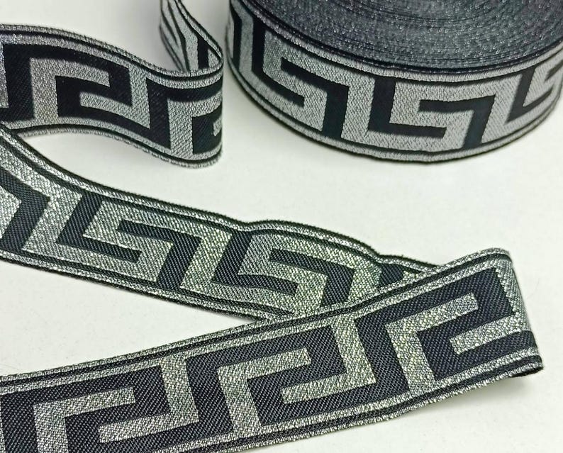 May include: Black and silver decorative ribbon with a Greek key pattern. The ribbon is rolled and unrolled, showcasing the repeating geometric design. The ribbon's metallic sheen adds a touch of elegance to the overall appearance.